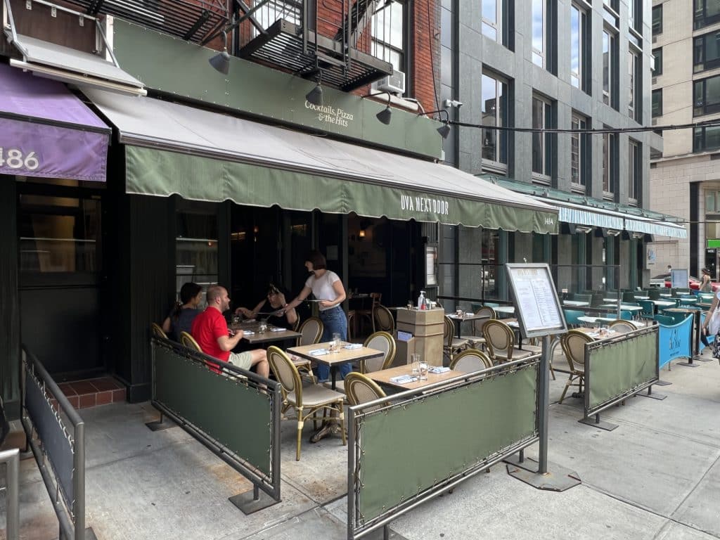 Photo shows a restaurant storefront with a green roll-out awning reading "UVA next door." Tables and chairs set up on the sidewalk are surrounded by barriers colored the same green.