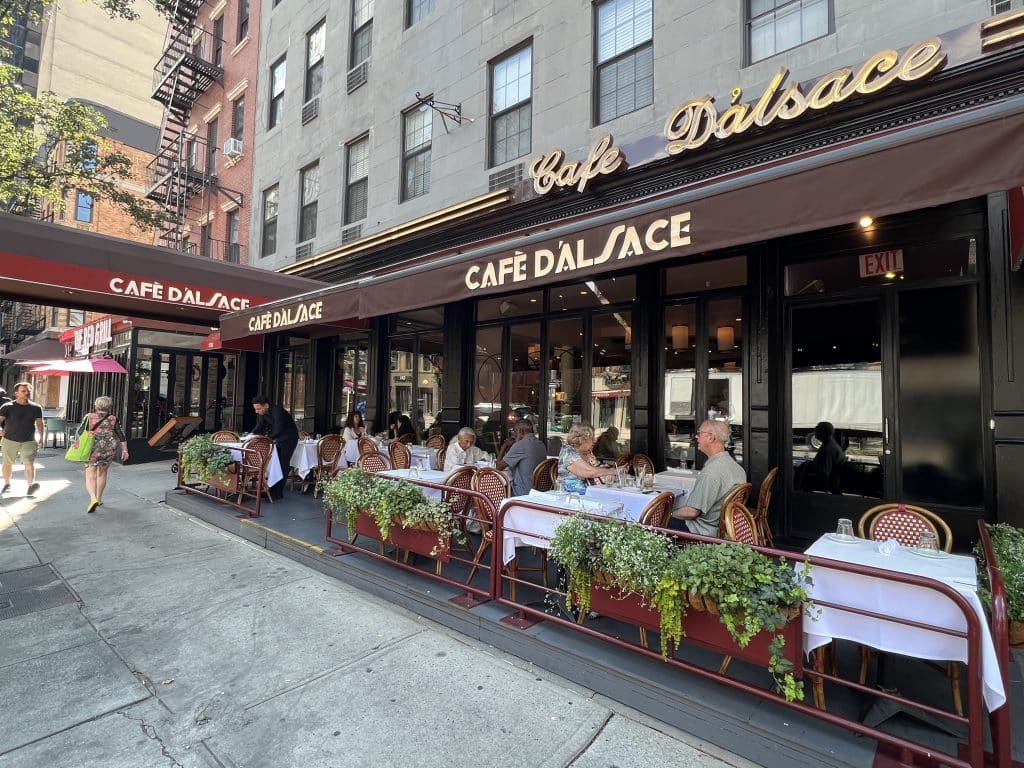Photo shows an angled view of outdoor dining in front of a restaurant with brown roll-out awnings reading 'Cafe D'Alsace.'