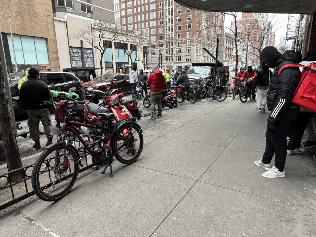 Delivery workers crowd the sidewalk and street with ebikes and moped | Upper East Site