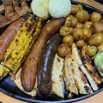 Photo shows a black plate with fried pork skin, sausages, roasted corn, a deep fried plantain, small roasted potatoes, grilled chicken and guacamole.