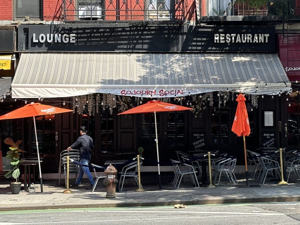 Photo shows a black storefront with a grey roll-out awning with the name "Sojourn Social" written in red. Tables are stationed in front on the sidewalk with red outdoor umbrellas providing shade.