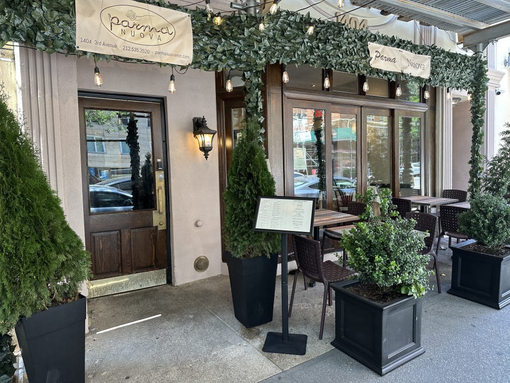 Photo shows a beige restaurant entrance with fake foliage adorning the top. Multiple plants are located in front.