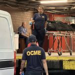 Photo shows a woman dressed in dark blue from the Medical Examiner's Office standing on a loading dock holding a gurney carrying a body in a black plastic bag, pointing to the back of a white van with a black horizontal stripe. Her partner, whose back of shirt reads 'OCME' in large yellow letters, is seen from behind looking up at the woman.