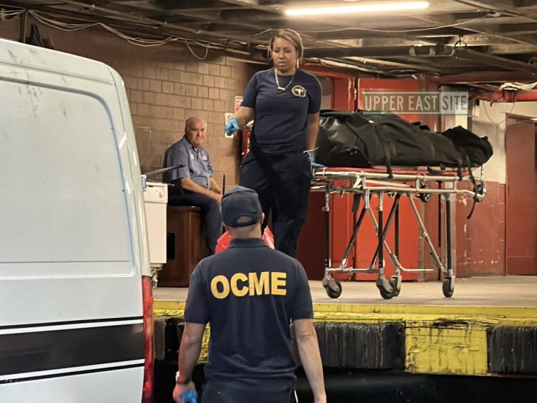 Photo shows a woman dressed in dark blue from the Medical Examiner's Office standing on a loading dock holding a gurney carrying a body in a black plastic bag, pointing to the back of a white van with a black horizontal stripe. Her partner, whose back of shirt reads 'OCME' in large yellow letters, is seen from behind looking up at the woman.