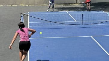Photo shows three women on a blue pickleball court with one of them hitting the ball with their paddle.
