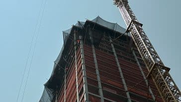 Photo shows a high rise building under construction with a tower crane high above it as seen from the ground looking upward.