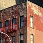 Photo shows a the top 1.5 stories of a red brick apartment building in a state of disrepair with a patchwork of facade repairs.