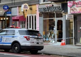 Photo shows a marked NYPD cruiser with its emergency lights on parked in front of a liquor store with a shattered glass door wrapped with police tape