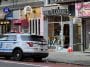 Photo shows a marked NYPD cruiser with its emergency lights on parked in front of a liquor store with a shattered glass door wrapped with police tape