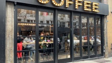 Photo shows an all glass storefront with people sitting drinking coffee behind the windows and a large sign on top that reads COFFEE in lights