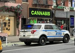 Photo taken from an angle looking shows an NYPD SUV parked in front of an illegal weed shop with a large black sign with lime green sign reading CANNABIS in all capital letters.