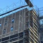 Photo shows a three-quarter view of the top two floors of a brown brick apartment building wrapped in scaffolding
