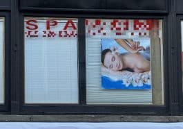 Photo shows two windows will the blinds drawn shut. Lettering on the window reads SPA and lists a website and a phone number twice, which have been blurred. A large photo in the window shows a woman laying on her stomach with hands massaging her back.