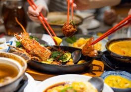 Photo shows a table with several Chinese food dishes and a plate of lobster in the center with three hands using chop sticks to grab a piece