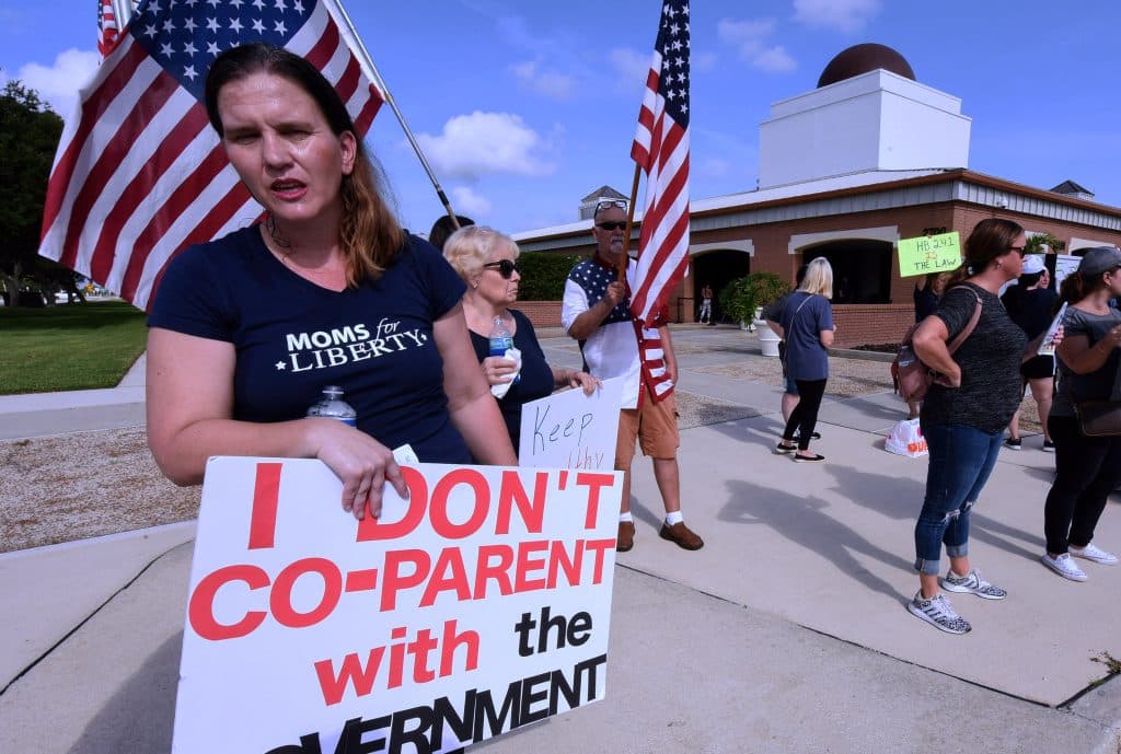 Photo shows a woman in a shirt reading 'Moms for Liberty' holding a sign reading 'I don't co-parent with the government.' Other protesters in the background wearing the same shirt are carrying large American flags 