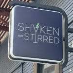 Photo shows dark blue square sign attached to a building by a black fixture and featuring the logo for 'Shaken Not Stirred.'
