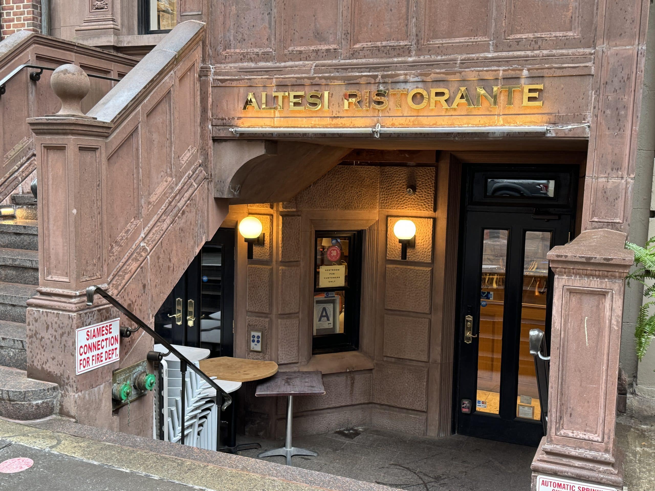Photo shows a staircase leading down front he sidewalk to the lower level of a building with a glass doors at the booth. A sign above the entrance reads "ALTESI RISTORANTE."