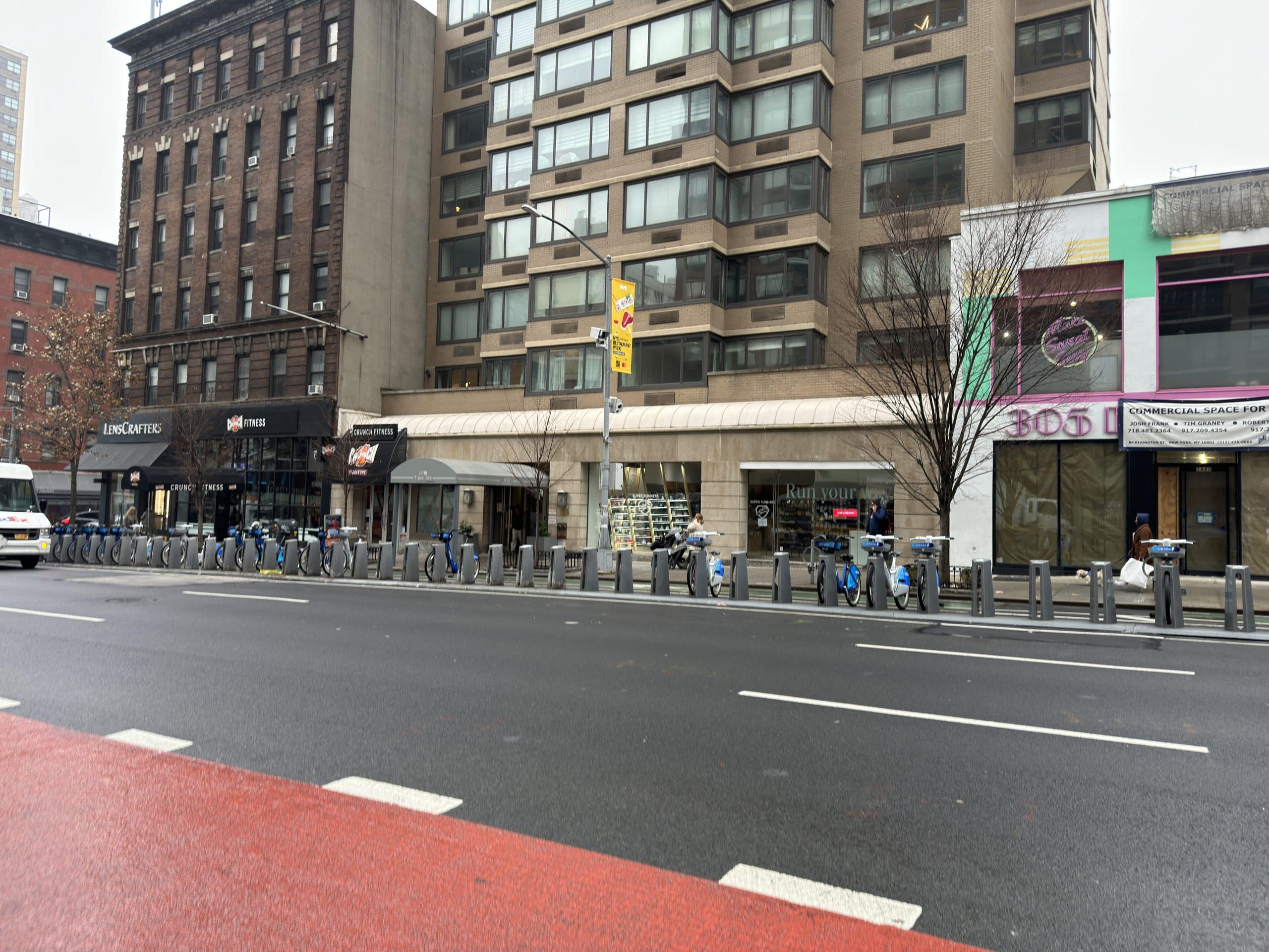 Photo shows a wide shot a row of more than three dozen bike docks lined up creating an obstacle in front of a beige apartment building whose awning extends to the curb.