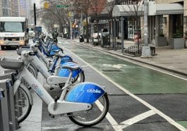 Photo shows a row of dozens of Citi Bike e-bikes, extra-wide bike lane and the sidewalk in front of a building where the awning extends to the curb.
