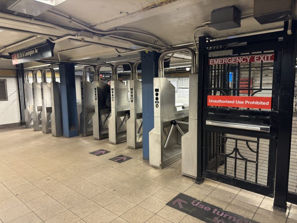 Photo shows a row of turnstiles and a black emergency exit door inside a NYC subway station.
