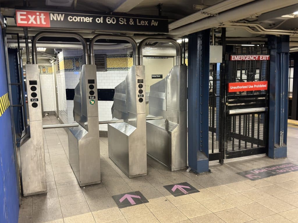 Photo shows a short row of turnstiles and a black emergency exit door inside a NYC subway station.