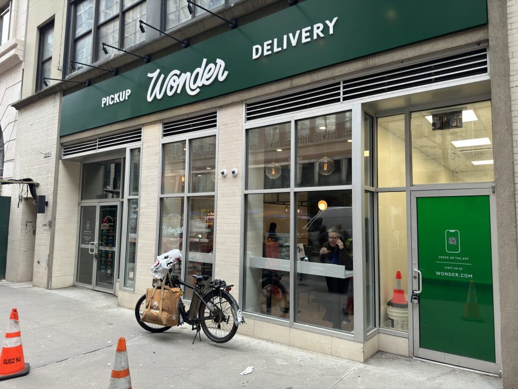 Photo shows an angled view of a beige brick and glass storefront with a green sign featuring white writing reading 'Wonder' and 'Pickup' and 'Delivery.' A bike is parked in front.