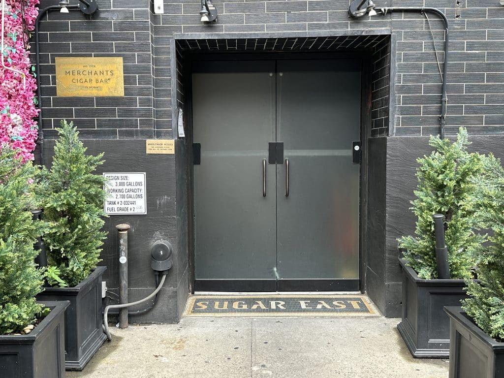 Photo shows the entrance to a black brick building with frosted glass doors. A small bronze sign on the left reads "Merchant's Cigar Bar' while small shrubbery lines the entranceway.