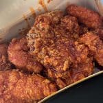 Photo shows a paper box filled with fried chicken tenders coated in a dark reddish-brown sauce