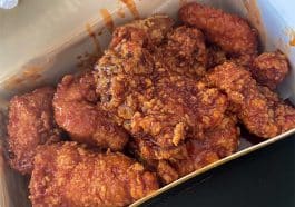 Photo shows a paper box filled with fried chicken tenders coated in a dark reddish-brown sauce