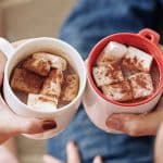 Photo shows a man's and woman's hands holding white mugs with hot chocolate and marshmallows.
