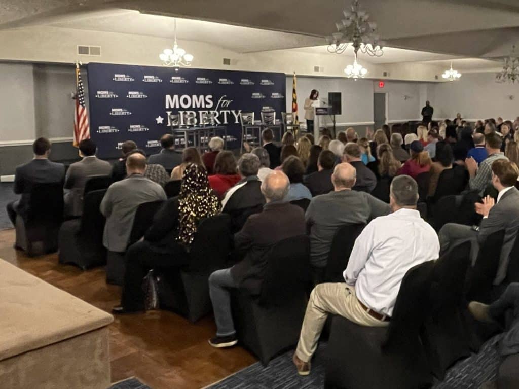 Photo shows a large conference room filled with seated guests with a stage in the background featuring a large 'Moms for Liberty' backdrop.