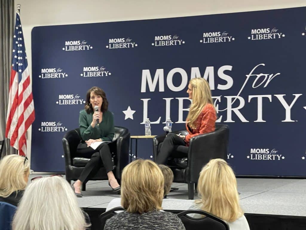 Photo shows two white women, a brunette and a blonde, on a stage with a large dark blue 'Moms for Liberty' backdrop behind them 