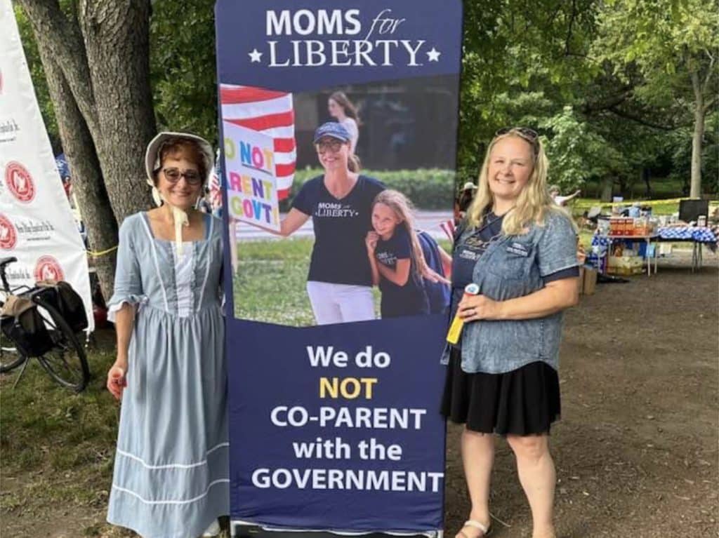 Photo shows two women, one in light blue Revolutionary War-era dress, standing next to a banner for Moms for Liberty which states 'We do NOT CO-PARENT with the GOVERNMENT'  