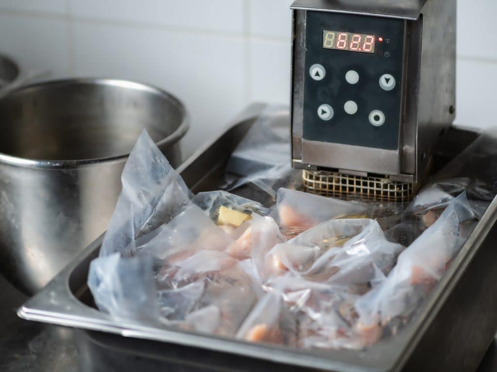 Photo shows a sous vide water warmer in a metal basin with several sealed bags full of chicken.