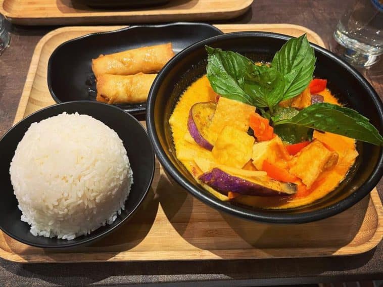 Photo show a wooden tray with three black dishes on it. One is a bowl filled with a yellow and red curry dish topped with fresh basil leaves. the Second is a dome-shaped scoop of white rice. While the third plate features two deep fried spring rolls.