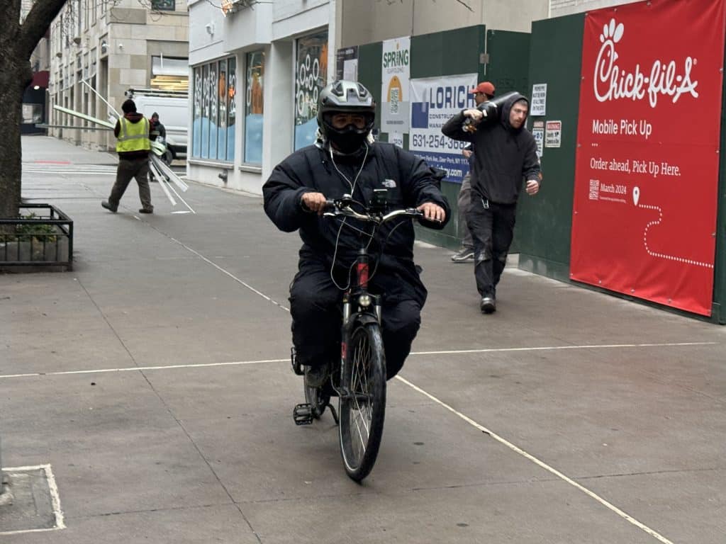 Photo shows a delivery worker in a motorcycle helmet riding an electric bike on the sidewalk.