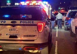 Photo shows the rear of a parked NYPD SUV with an ambulance in front of it, both with emergency lights on, and NYPD officers standing between them and a parked row of cars on the right.