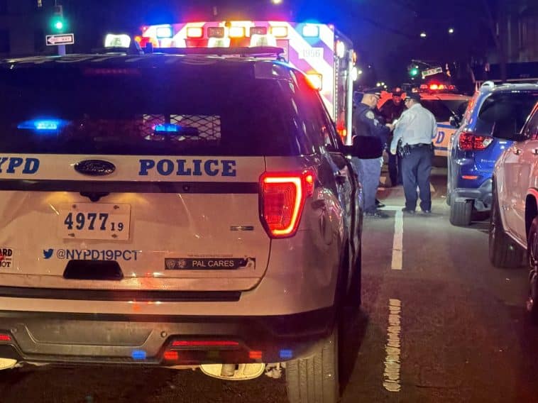 Photo shows the rear of a parked NYPD SUV with an ambulance in front of it, both with emergency lights on, and NYPD officers standing between them and a parked row of cars on the right.