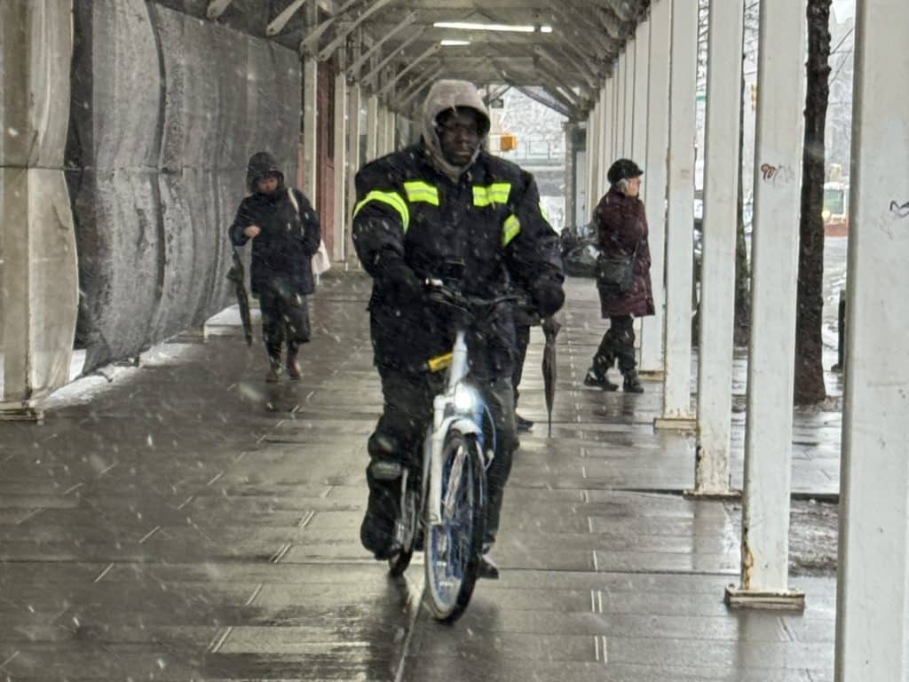 Photo shows a delivery workers riding an electric bike on a sidewalk in the snow.