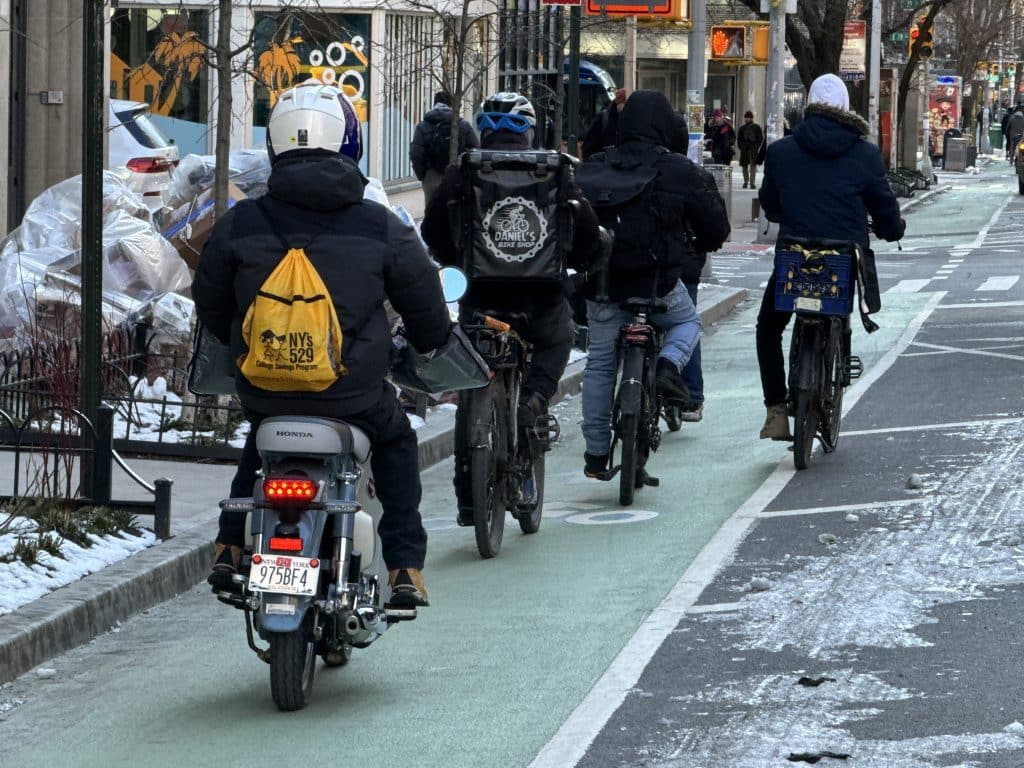 Photo shows a person on a moped illegally riding with a group of delivery workers on e-bikes.