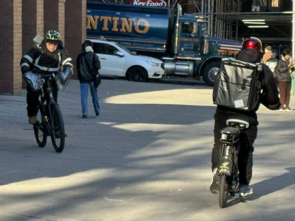 Photo shows two delivery workers riding an electric bike on a sidewalk.
