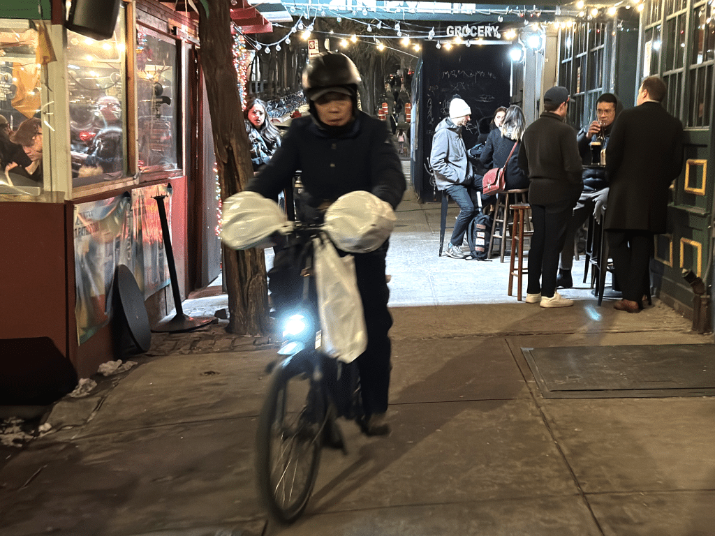 Photo shows a delivery worker on an electric bike riding on the sidewalk through a bars outdoor dining setup.