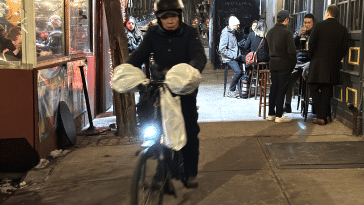 Photo shows a delivery worker on an electric bike riding on the sidewalk through a bars outdoor dining setup.