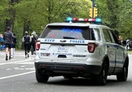 Photo shows an NYPD cruiser with its emergency lights on in Central Park with joggers running nearby.