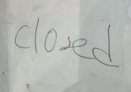 Photo shows a handwritten sign made from a white sheet of paper behind glass door reading, 'closed.'