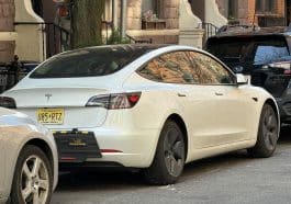 Photo shows a white Tesla with New Jersey plates parked on a residential side street.