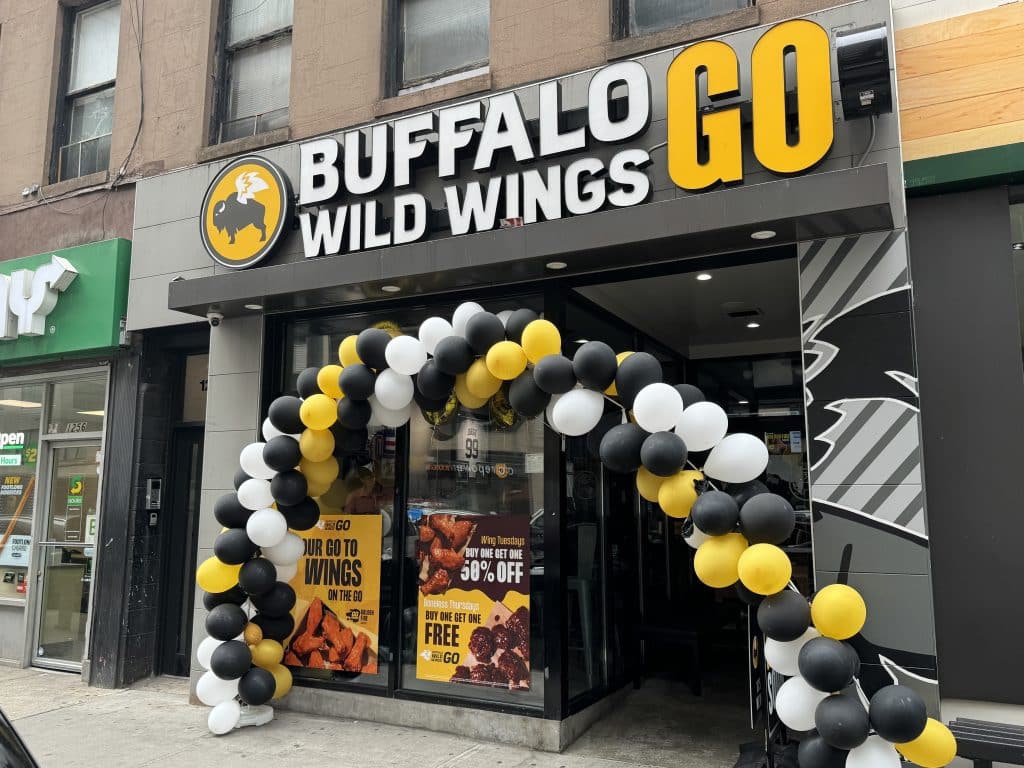 Photo shows a grey storefront featuring a sign reading 'Buffalo Wild Wings GO' and a black yellow and white balloon arch in front.