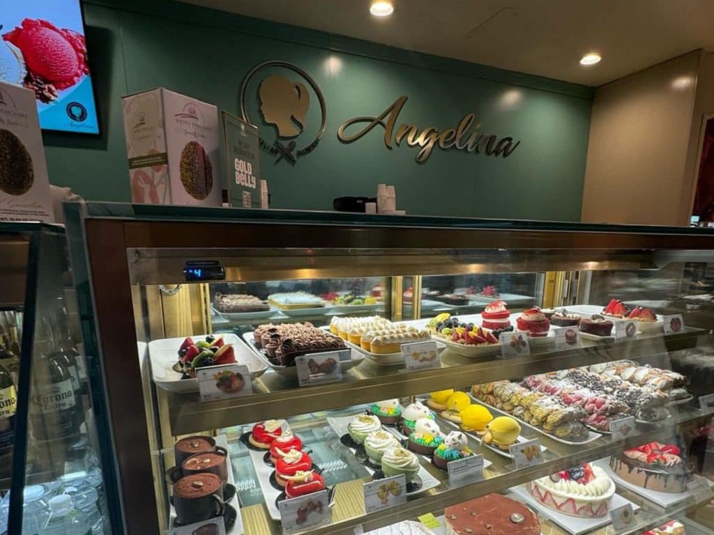 Photo shows a display case full of cakes and pastries with a green wall behind it and a script logo reading ‘Angelina.’