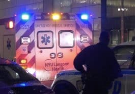 Photo shows a dark silhouette of a cop standing behind an ambulance with its emergency lights on at night.
